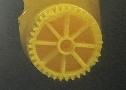 Yellow plastic gear with toothed edge and radial spokes, likely for ID card printer mechanism